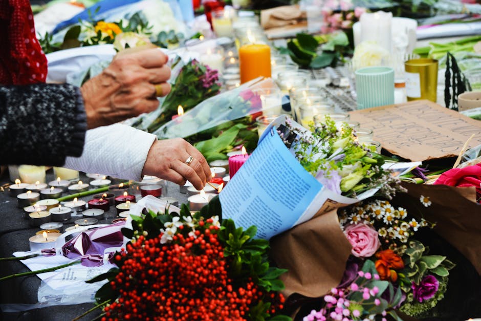 A person arranging floral tributes at a memorial, with multiple bouquets of fresh flowers including roses, daisies, and lilies in various colors such as pink, white, purple, and orange. The arrangemen