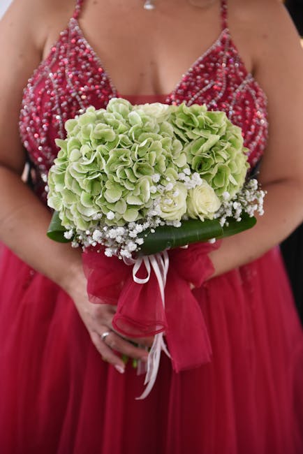 A person dressed in a vibrant red gown, adorned with intricate beaded embellishments on the bodice, holds a round floral bouquet comprising pale green hydrangeas with ruffled petals, interspersed with