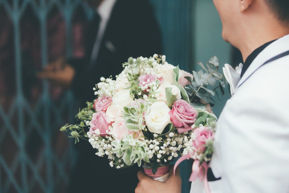A person in a white suit, possibly a groom, is holding a round floral bouquet comprising pink roses, white roses, and white baby's breath, accented with green leaves and foliage. The bouquet is wrappe