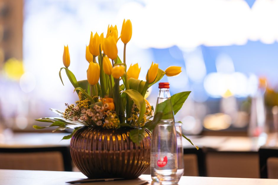 A floral arrangement featuring bright yellow tulips with elongated, smooth petals standing upright in a round, dark brown glass vase with vertical ridges. Surrounding the tulips are small sprigs of de