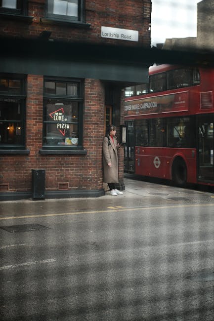 A person standing on a wet street corner in Haringey N8, dressed in a long beige coat and white sneakers, waits beside a red double-decker bus parked near a brick building with dark-framed windows. Th