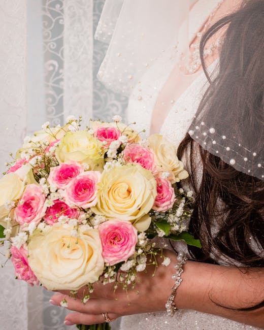 A bride holding a floral bouquet composed of large cream roses and smaller pink roses, accented with delicate white baby's breath. The bouquet is arranged in a rounded, compact style and wrapped with 