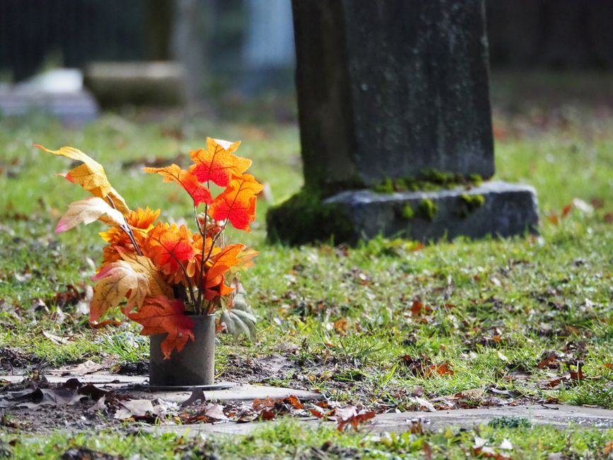 A floral tribute consisting of a bouquet of artificial autumn leaves in shades of orange, red, yellow, and cream arranged in a small, cylindrical metal container. The bouquet is positioned on the gras