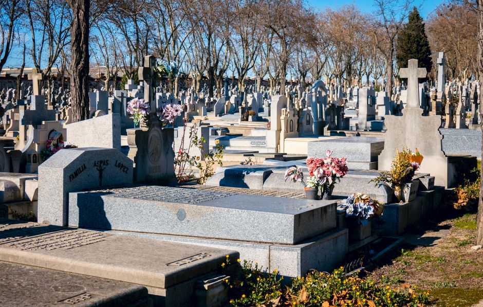 A view of a cemetery with numerous gray granite headstones and crosses, some decorated with fresh flower arrangements featuring pink, purple, and white blossoms. The flowers are placed in small black 